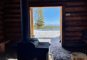 View from inside Meissner Warming Shelter showing a wood stove and snowy landscape with pine trees and a blue sky in the background, located at Virginia Meissner Sno-Park, Central Oregon.