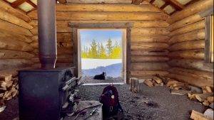 Inside a rustic log cabin at a Sno-Park, featuring a wood stove, firewood, a backpack, and a dog relaxing by the open doorway with a snowy landscape and trees in the background.