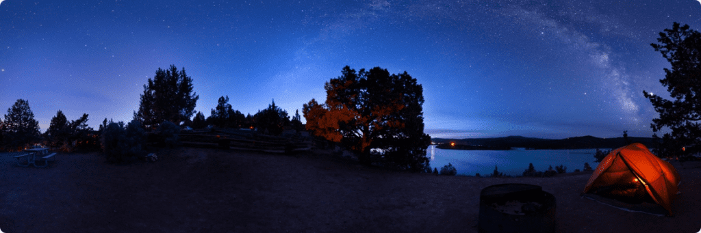 Campsite at night with a lit-up tent in the foreground and a view of lake and Milky Way in the background. Oregon State Parks, Prineville Reservoir State Park and International Dark Sky Park